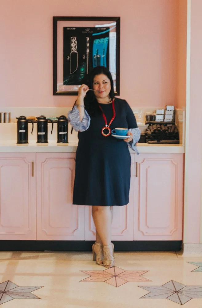 A woman with black hair in a black dress and striped sleeves holding a blue cup, standing in front of a pink counter with coffee dispensers, in a room with pink walls and star-patterned floor tiles.