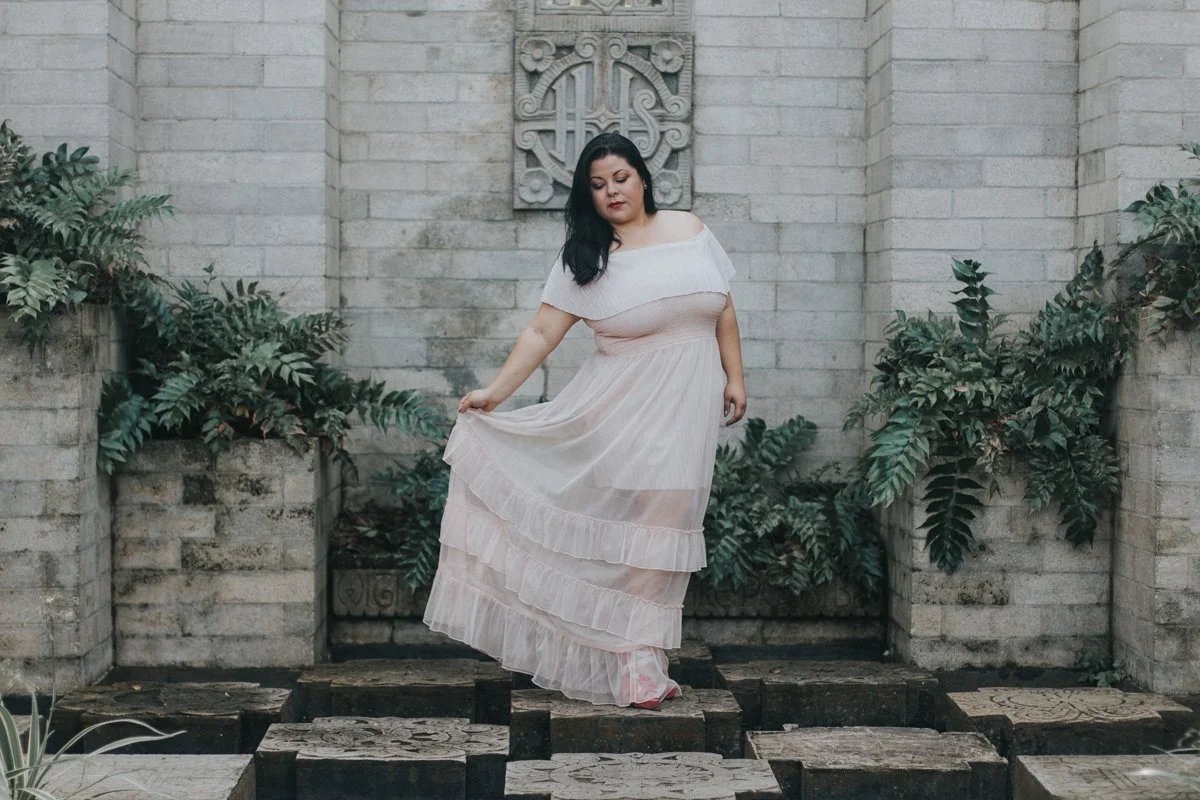 Woman in a beige dress standing on stone tiles among green plants and a brick wall background.