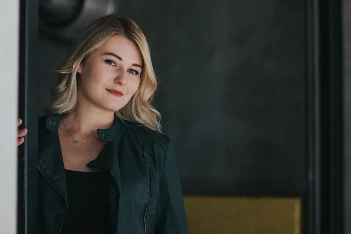 A young woman with blonde hair and blue eyes standing at a doorway, looking at the camera with a slight smile.