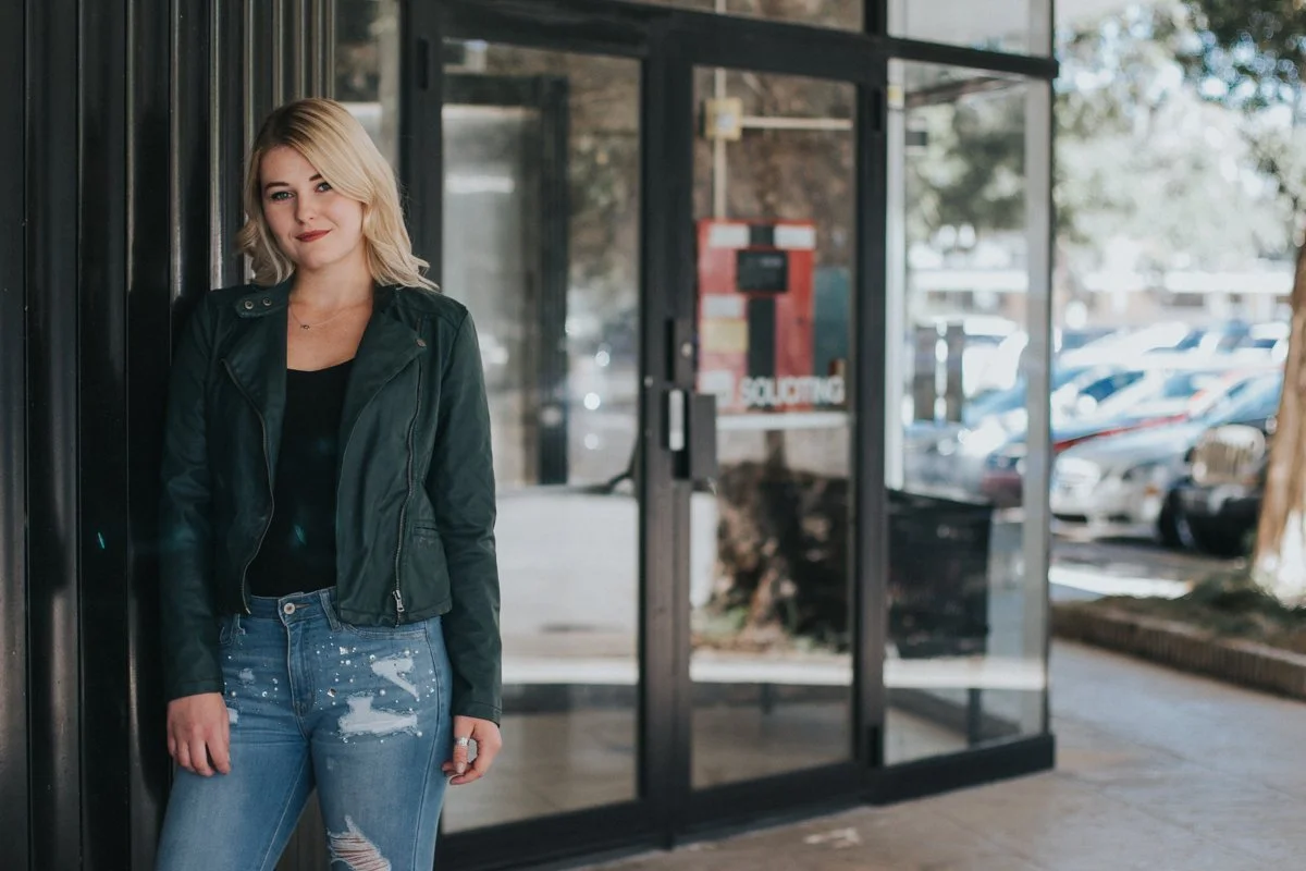 A young woman with blonde hair and a black leather jacket standing outside a storefront with a glass door and window, with a parking lot visible in the background.
