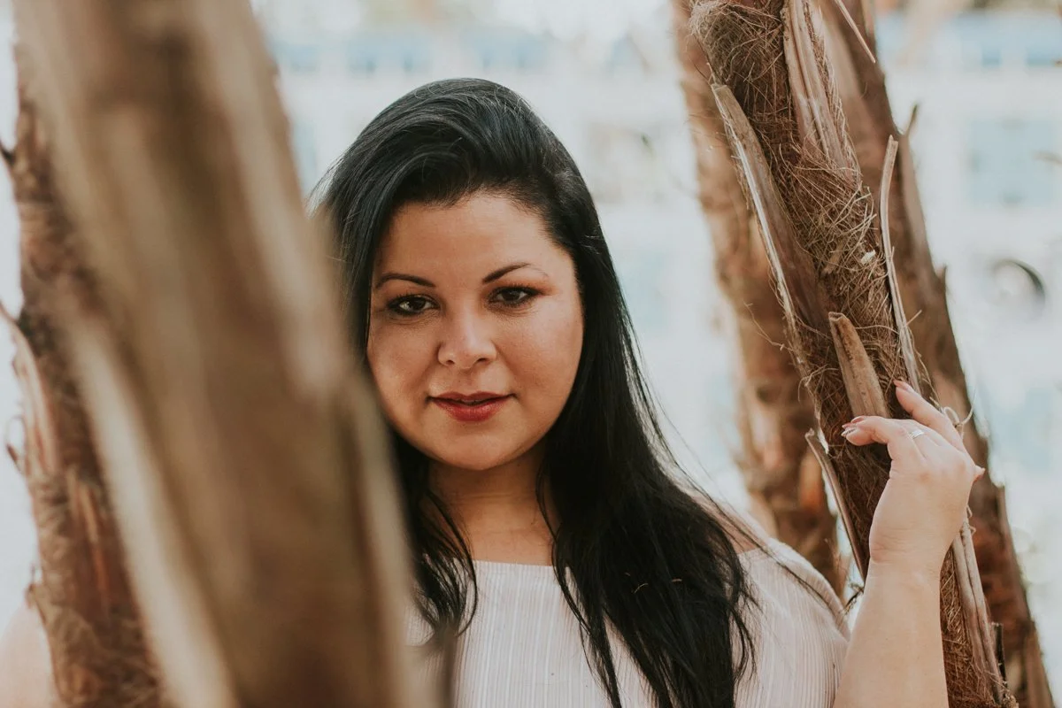 A woman with long black hair posing between trees or wooden poles outdoors.