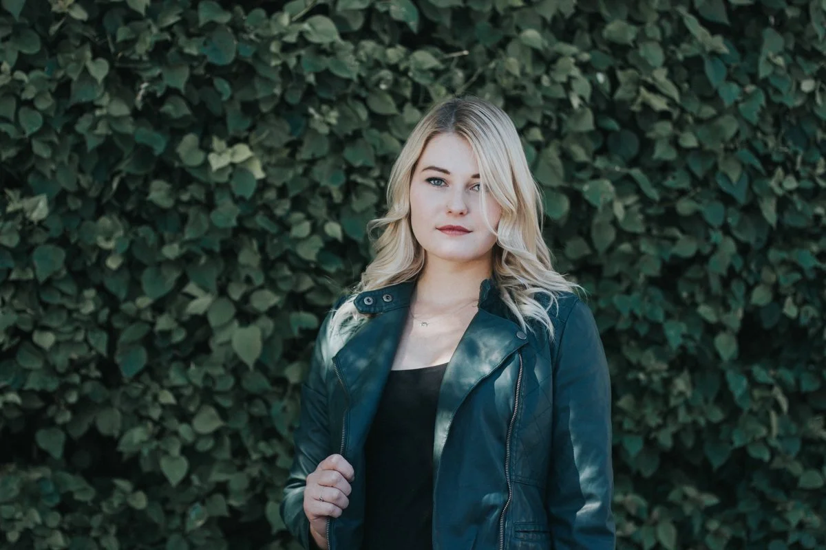 A young woman with blonde hair and a black leather jacket standing in front of a dense green leafy background.