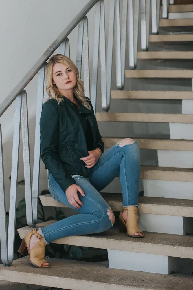 A woman with blonde hair sitting on stairs inside a building, looking at the camera, wearing a dark green jacket, ripped jeans, and tan high-heeled sandals.