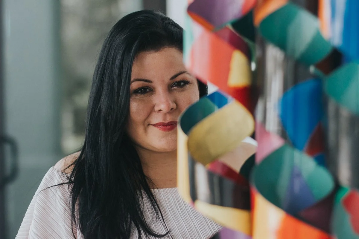 A woman with long black hair and light skin smiling at a colorful paper craft decoration.