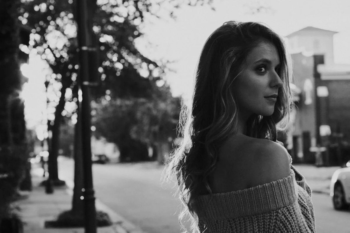 A young woman with wavy hair looking over her shoulder on a street at sunset, in black and white.