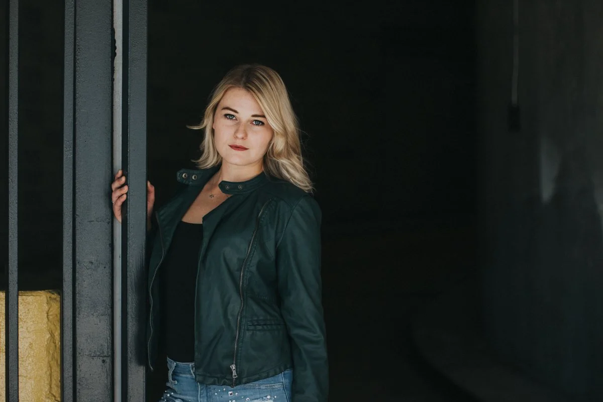 A young woman with blonde hair standing next to a dark gray wall, wearing a black shirt and a green jacket, looking at the camera.