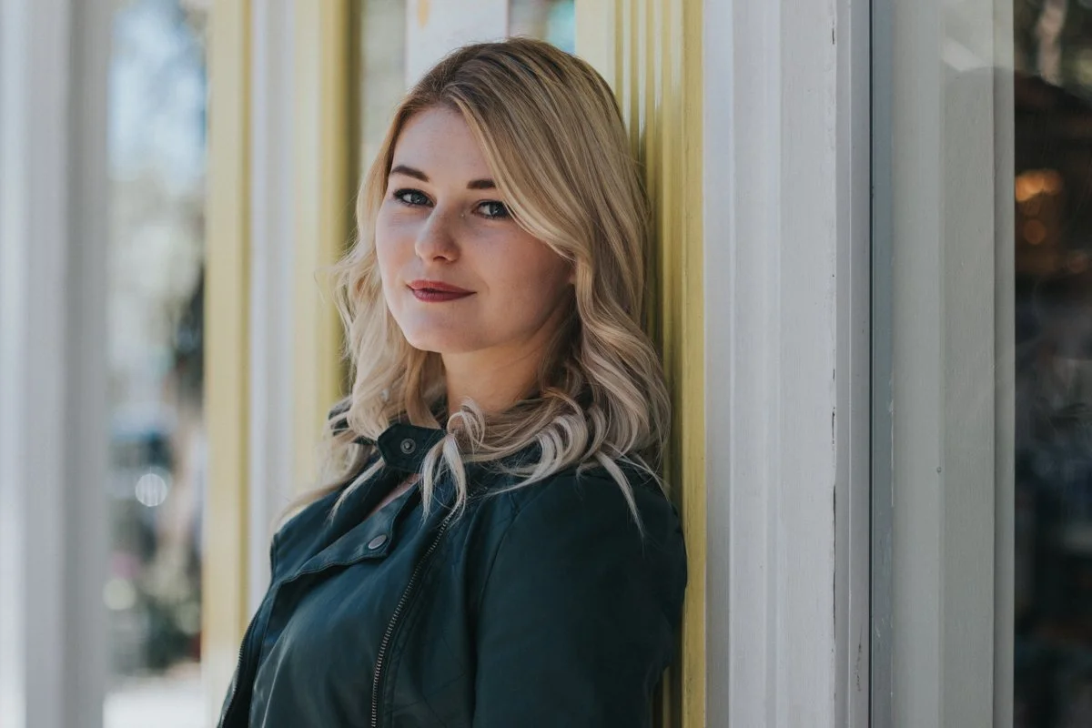 A young woman with blonde wavy hair and fair skin standing against a wall, wearing a black leather jacket, outdoors during daytime.