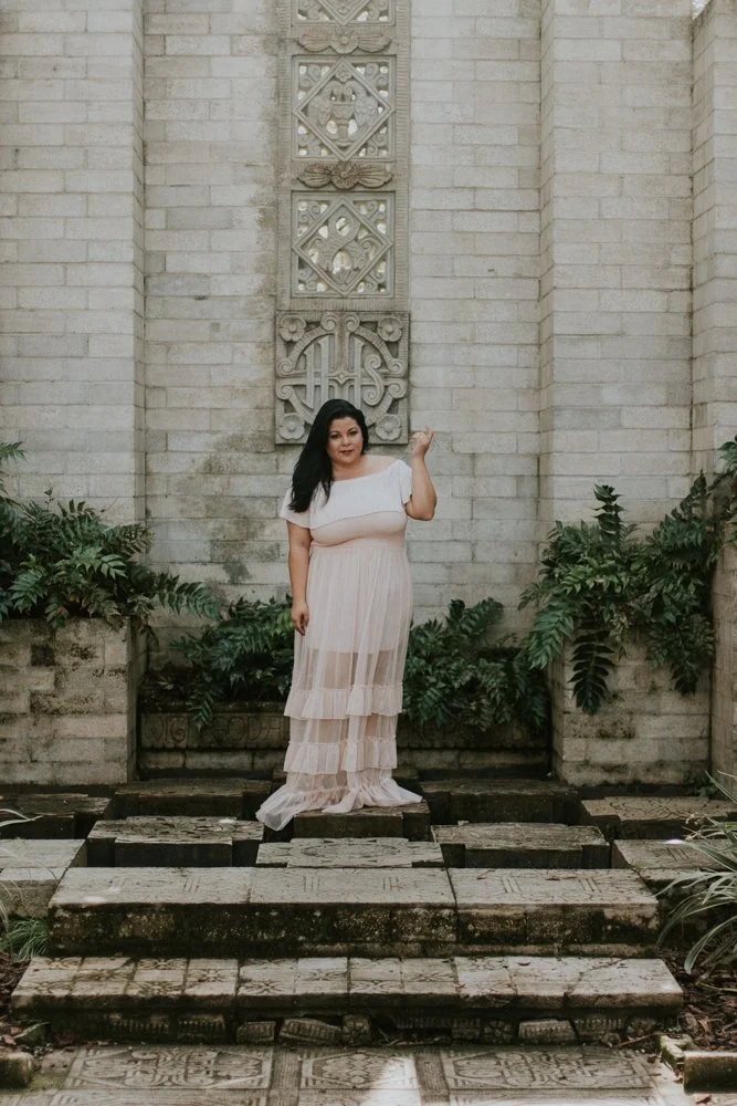 A woman in a long, light-colored dress standing on a stepped platform in front of a stone wall with decorative carvings, surrounded by lush green plants.