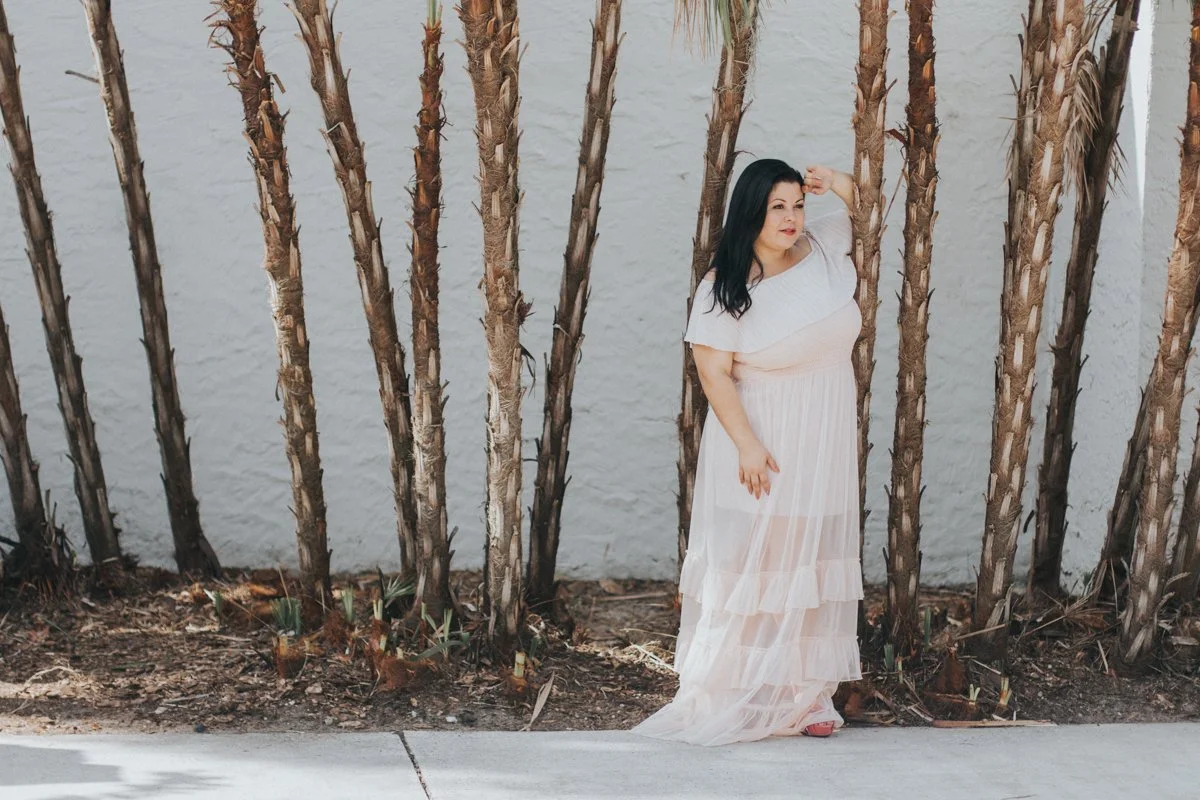 A woman with black hair wearing a white semi-sheer dress standing outdoors against a background of tall, thin palm trees and a white textured wall.