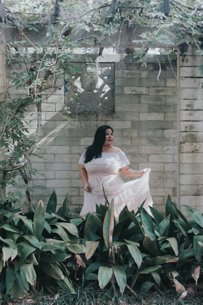 A woman in a light pink dress holding the hem in a garden with large green leaves, a brick wall, and a decorative metal artwork with a face above her.