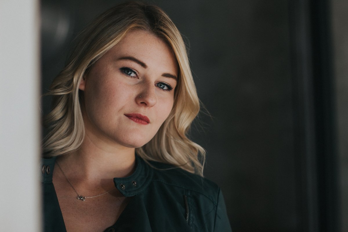 A young woman with blonde hair, blue eyes, and a nose piercing looks at the camera. She is wearing a dark green jacket and a delicate necklace, standing indoors near a window with a neutral background.