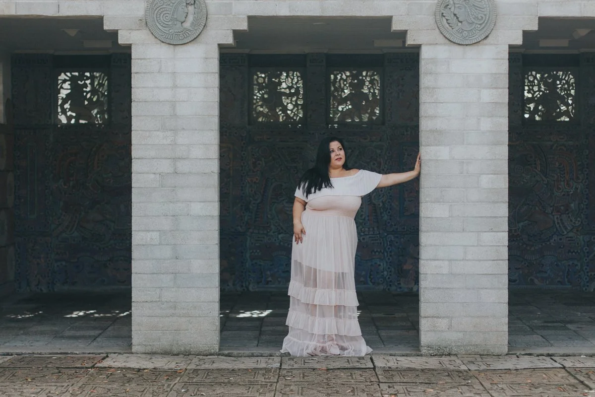 A woman in a long, tiered white dress standing under a stone archway with ancient carvings on the walls behind her, touching the pillar with her right hand.