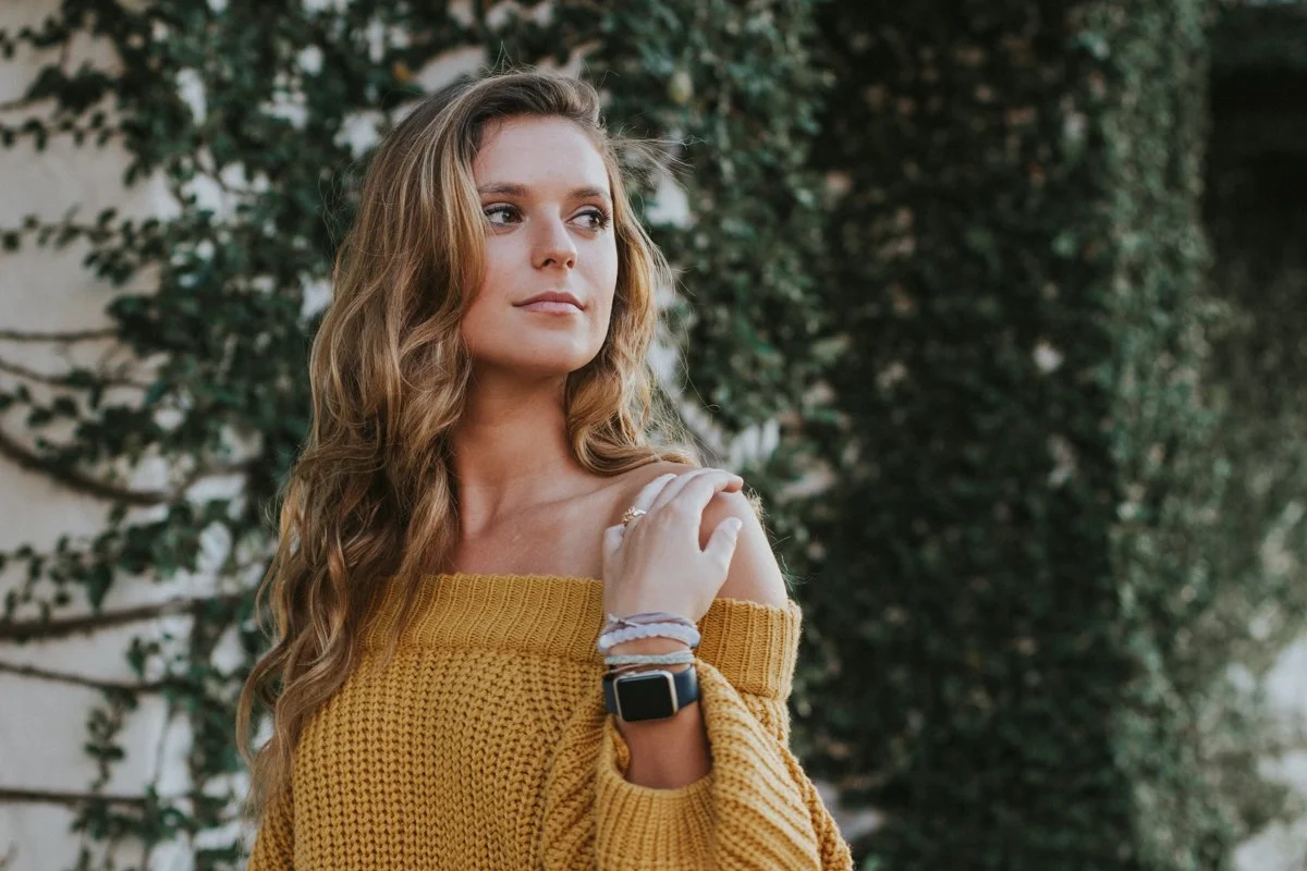 A woman with long wavy hair wearing a yellow off-shoulder sweater standing outdoors with green foliage in the background.
