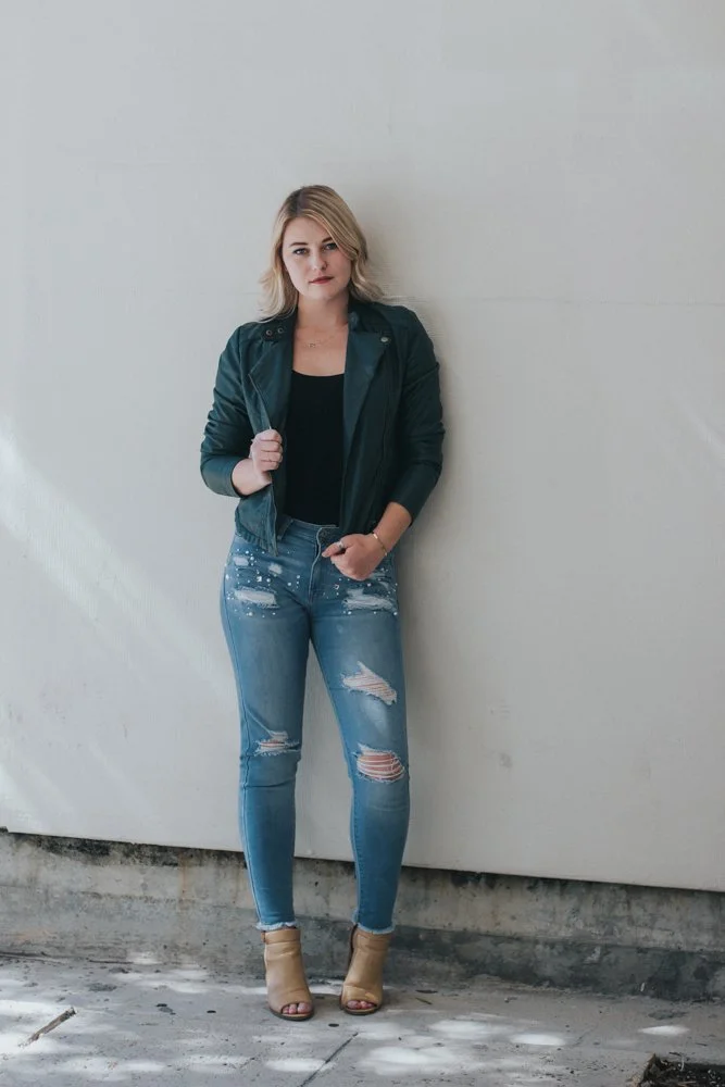 A young woman with blonde hair standing against a plain white wall, wearing a dark jacket, black top, ripped jeans, and open-toed tan high heels.