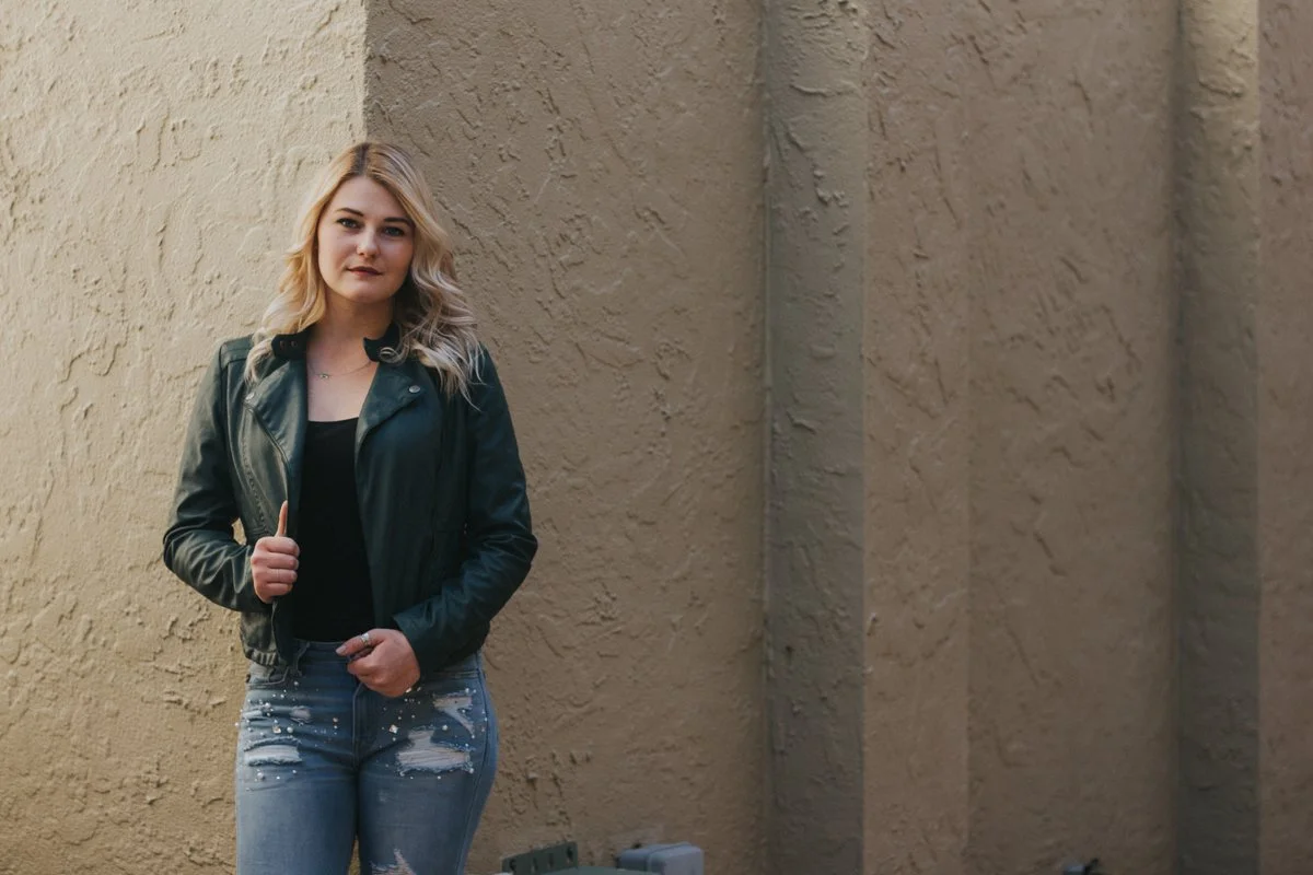 A young woman with blonde hair and fair skin stands against a beige textured wall, wearing a black leather jacket, black top, and distressed jeans with white paint splatters. She is looking directly at the camera with a neutral expression.