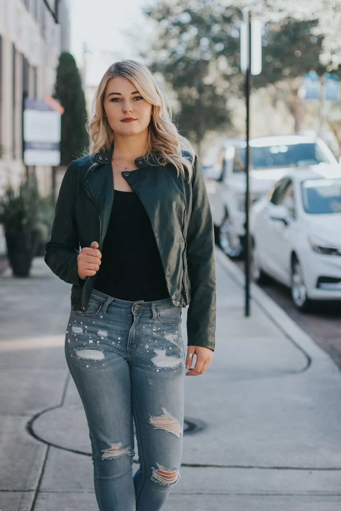 A young woman walking on a city sidewalk, wearing a black leather jacket and distressed jeans, with parked cars and trees in the background.