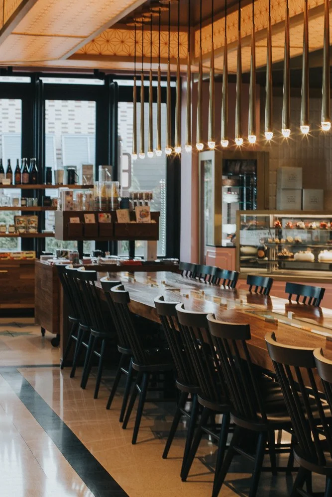 Interior of a modern cafe with high wooden tables, black chairs, hanging pendant lights, a display case with desserts, and shelves with bottles and products.