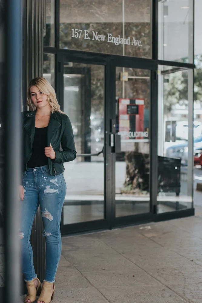 A young woman standing outside near the glass entrance of a building at 157 E. New England Ave, wearing a black top, black leather jacket, distressed blue jeans, and high-heeled shoes. She has blonde hair and is leaning slightly against a pole.