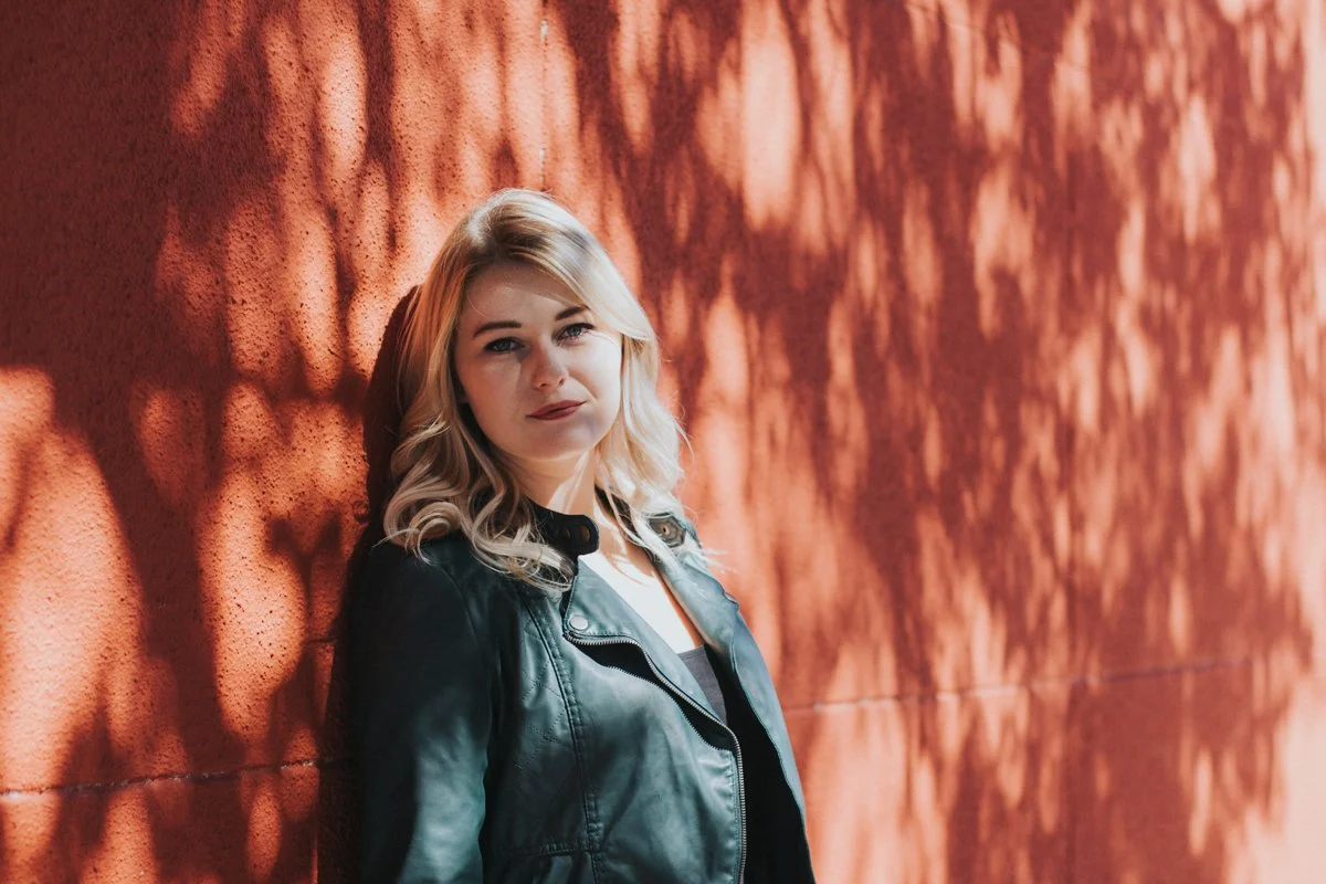 A woman with blonde wavy hair leaning against a red brick wall with dappled sunlight shadows, wearing a black leather jacket.