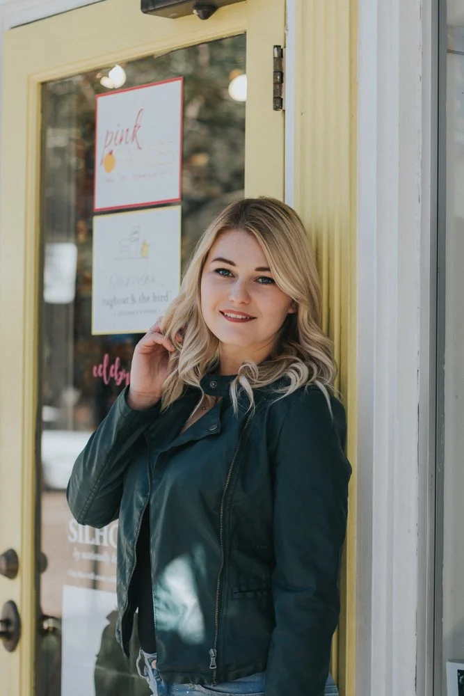A young woman with blonde hair, wearing a black leather jacket, leaning against a door with glass panels and yellow trim, smiling at the camera.