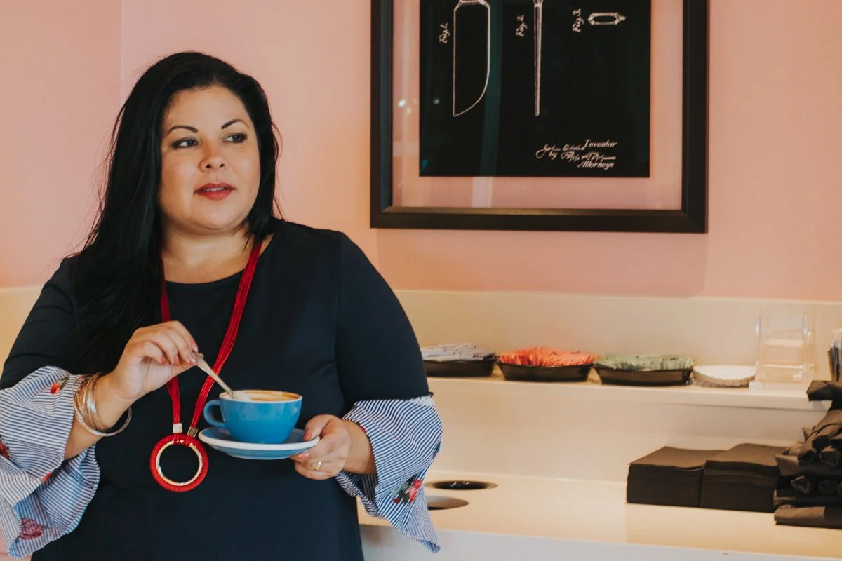 A woman with long black hair, wearing a black dress with striped sleeves and a red necklace, holding a blue cup and saucer, standing in a room with a pink wall, artwork on the wall, and stacked bowls in the background.