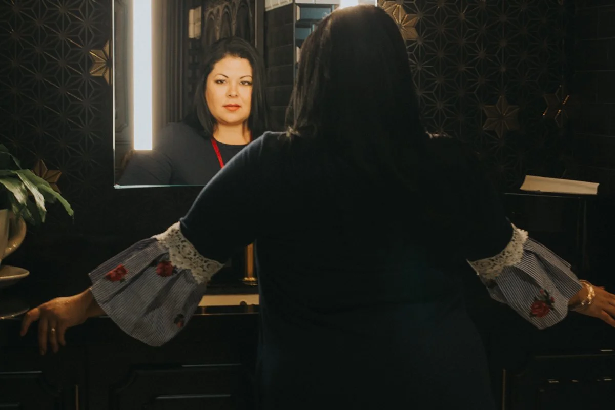 A woman with black hair and a black dress looking at her reflection in a mirror, with her arms resting on a piano, in a room with dark patterned walls and a potted plant.