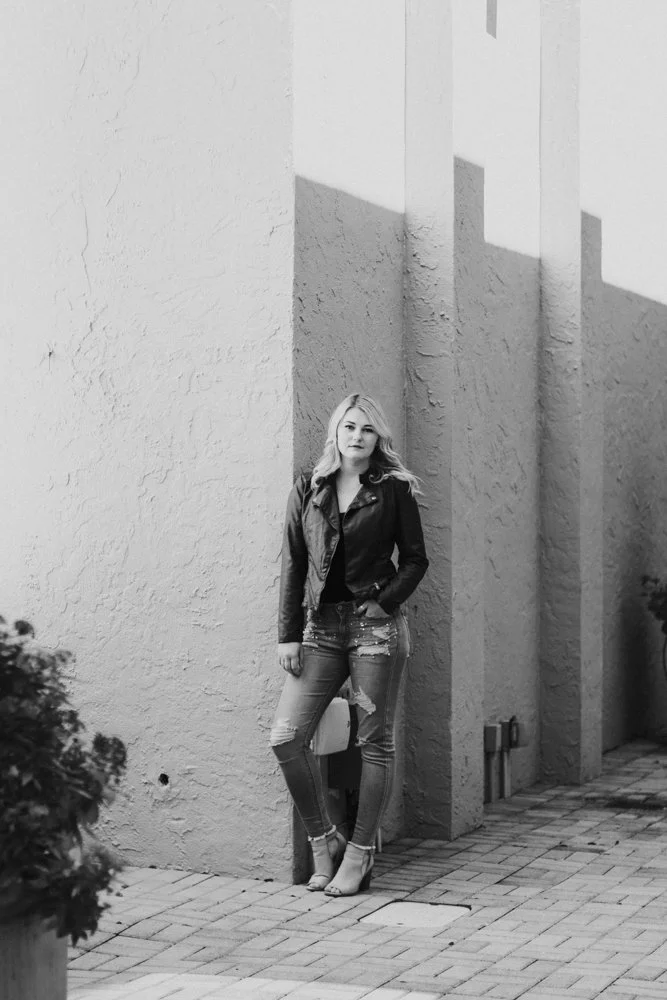 A woman with light-colored hair and light skin standing against a textured wall with geometric shadows in an outdoor setting. She is wearing a dark jacket, distressed jeans, and high-heeled shoes.