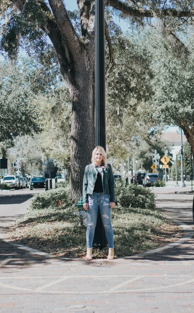 A woman standing on a sidewalk in front of a large tree and a pole, with cars and street signs in the background.