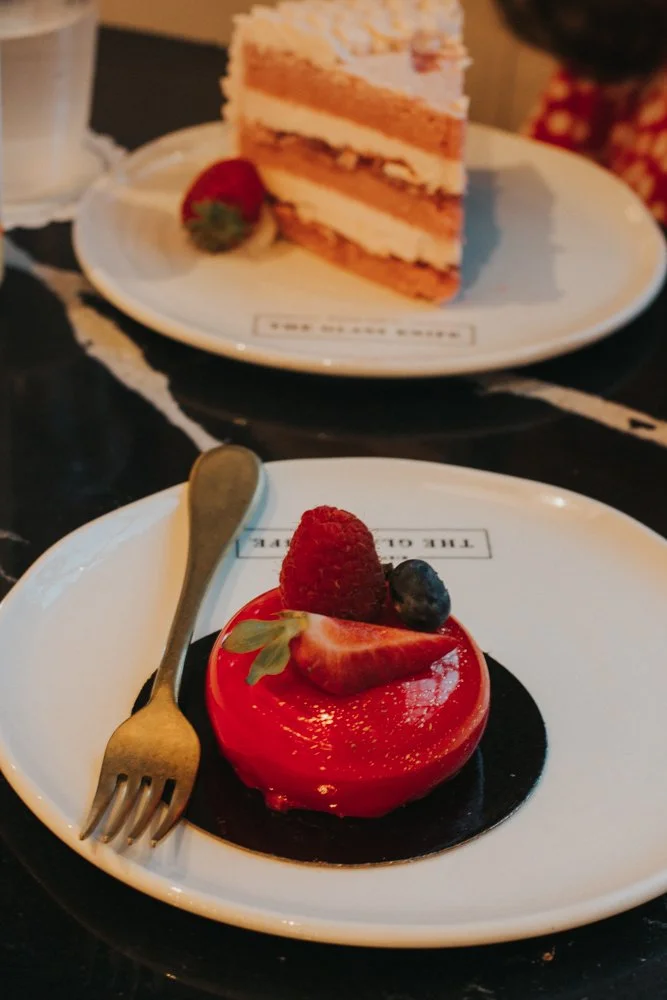 Close-up of a small, round, red glazed dessert topped with a strawberry, raspberry, and blueberry, served on a white plate with a gold spoon. In the background, a slice of layered strawberry cake with white frosting and a strawberry garnish is on ano