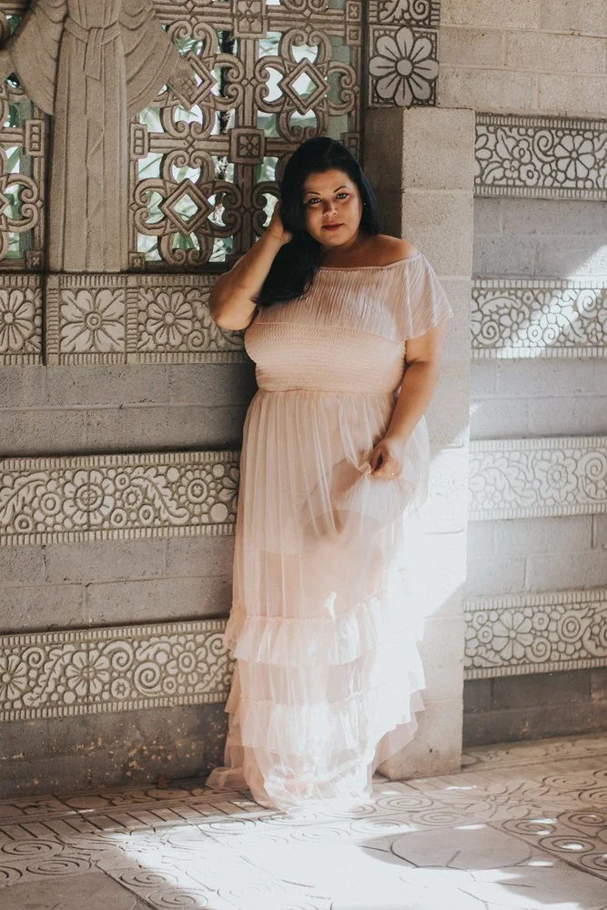 A woman in a light pink, off-the-shoulder dress standing against a decorative stone wall with intricate patterns and sculptures, with sunlight casting shadows on the ground.