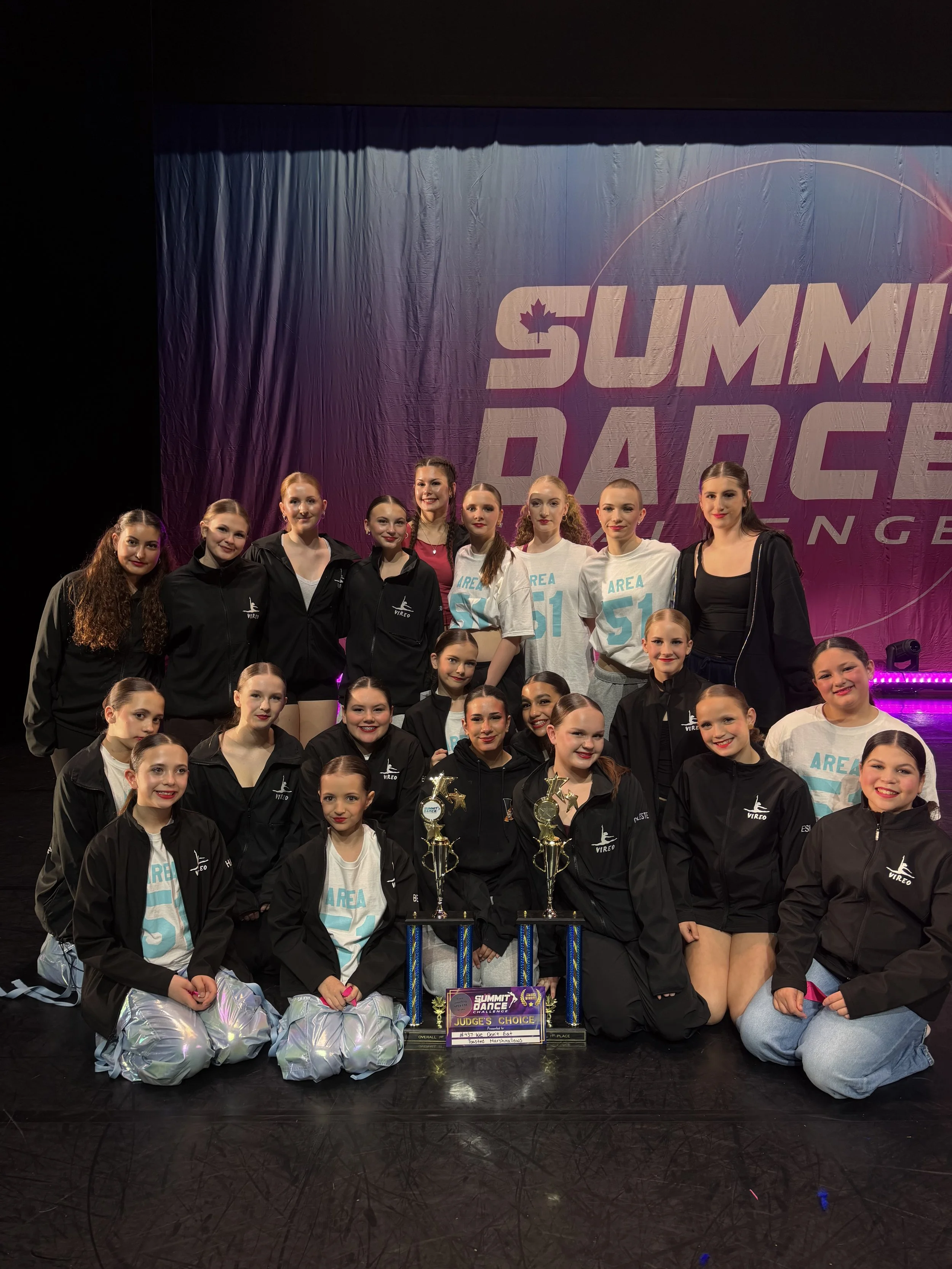 A group of young female dancers on stage holding awards and certificates, some kneeling and some standing, with a large screen behind them displaying the words "SUMMIT DANCE" and other promotional text.