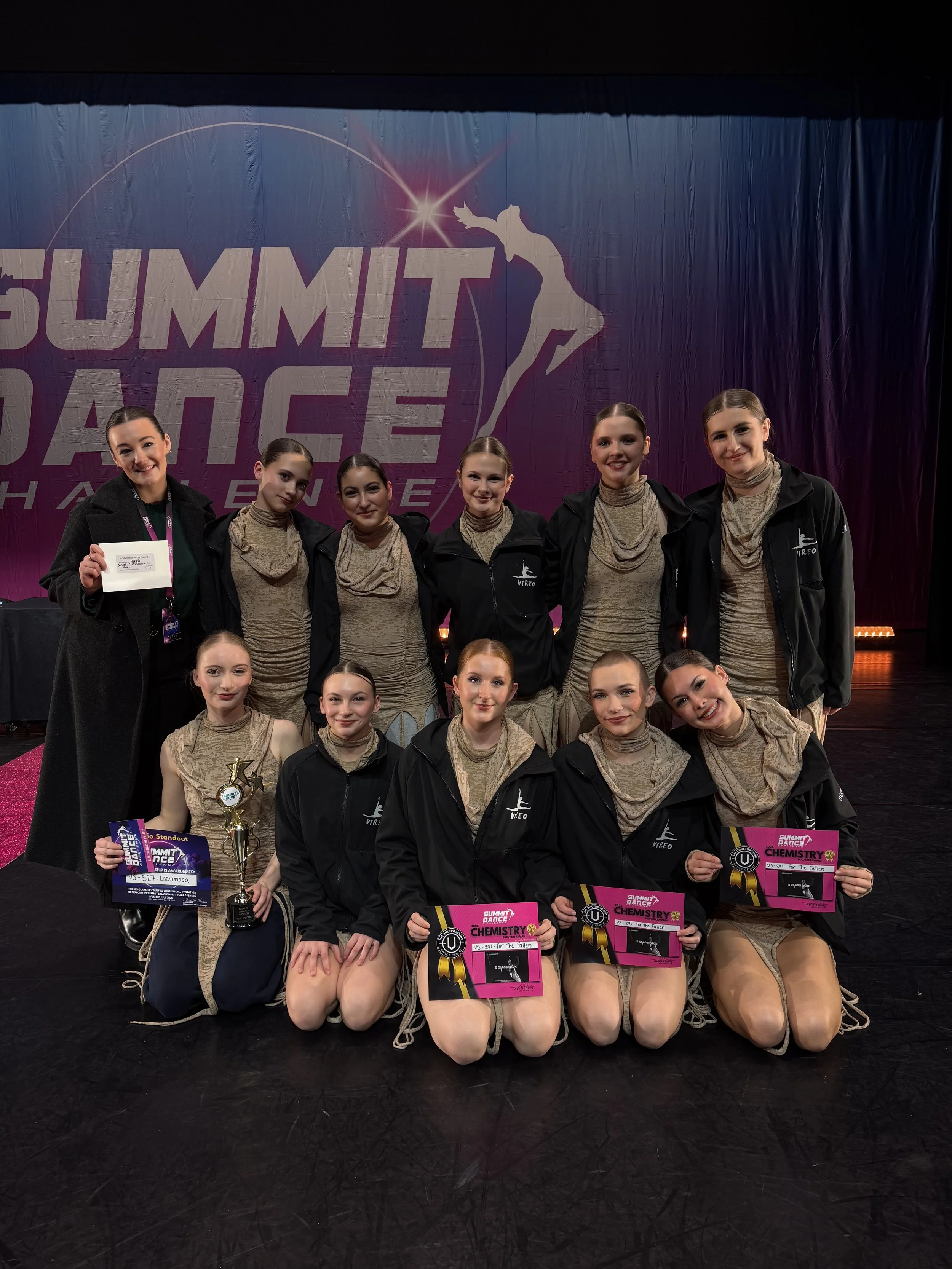 A group of young female dancers on stage holding awards and certificates, some kneeling and some standing on a dance stage.