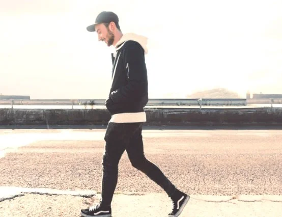 A man walking outdoors on a sunny day, wearing a black baseball cap, black hoodie with white lining, black pants, and black sneakers, with a cityscape in the background.