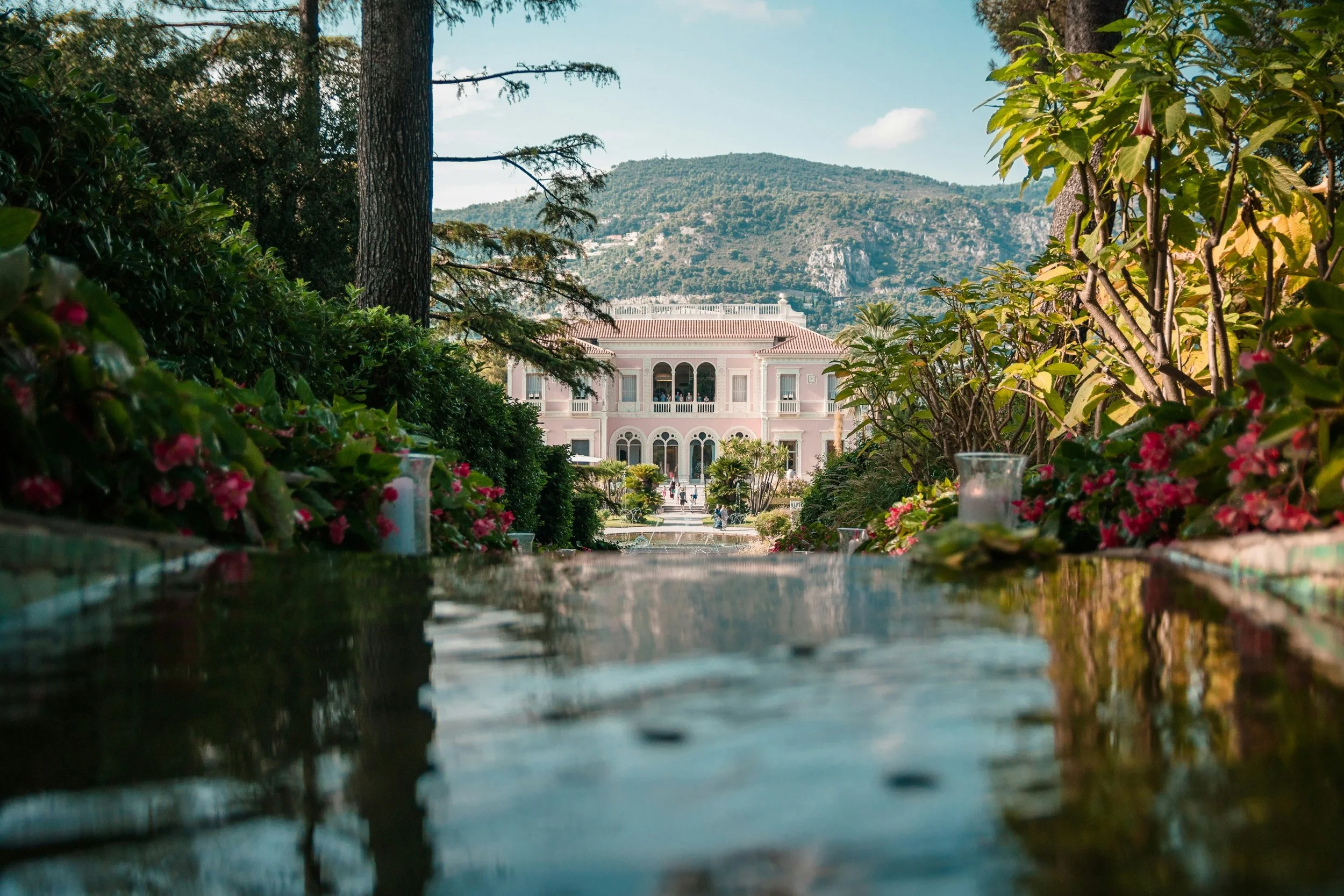 A landscaped garden leading to a grand, pink mansion with arched windows, overlooking a mountain in the background.