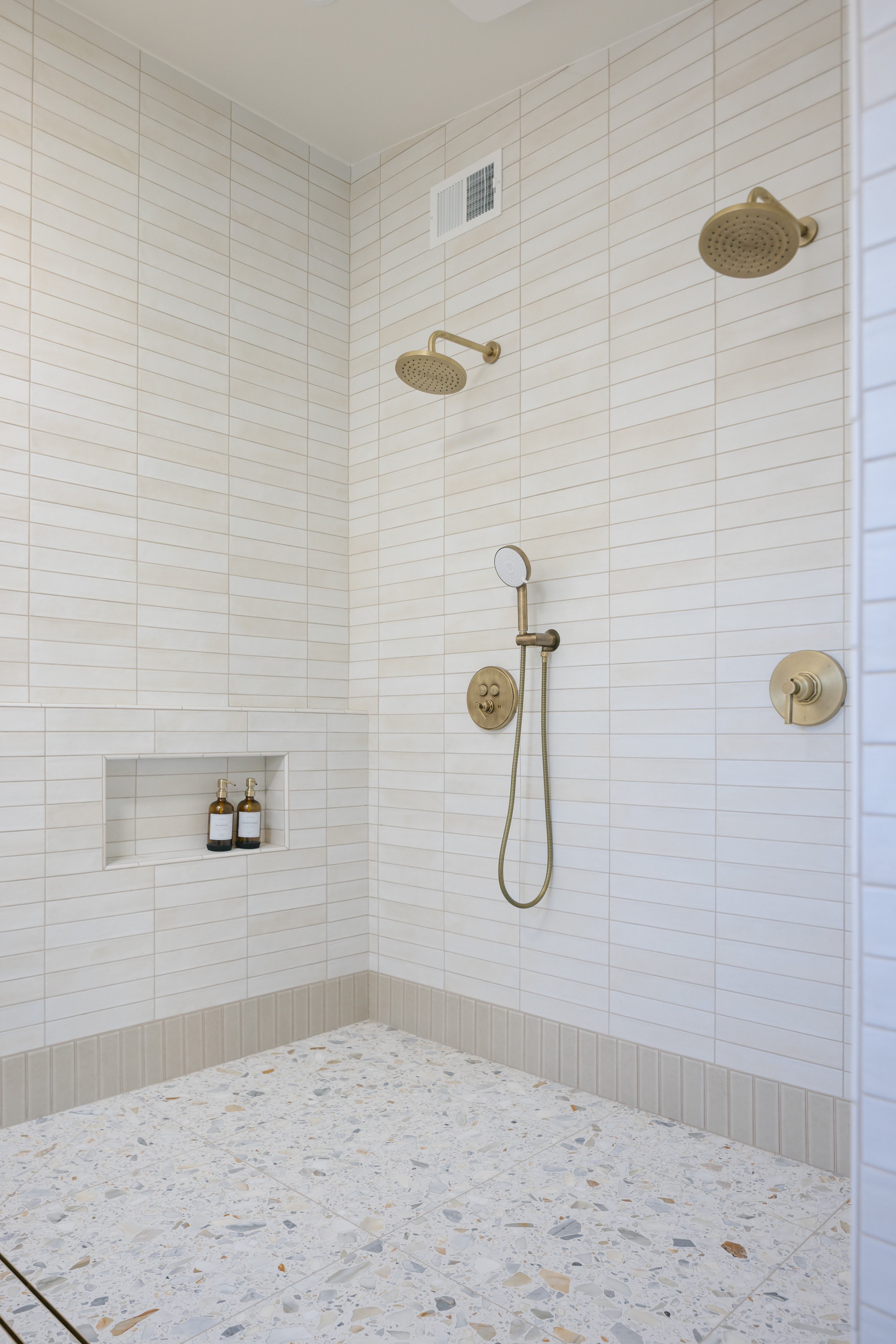 A modern walk-in shower with beige tile walls, two rainfall showerheads, a handheld shower, and a built-in shelf with two bottles of shower products.
