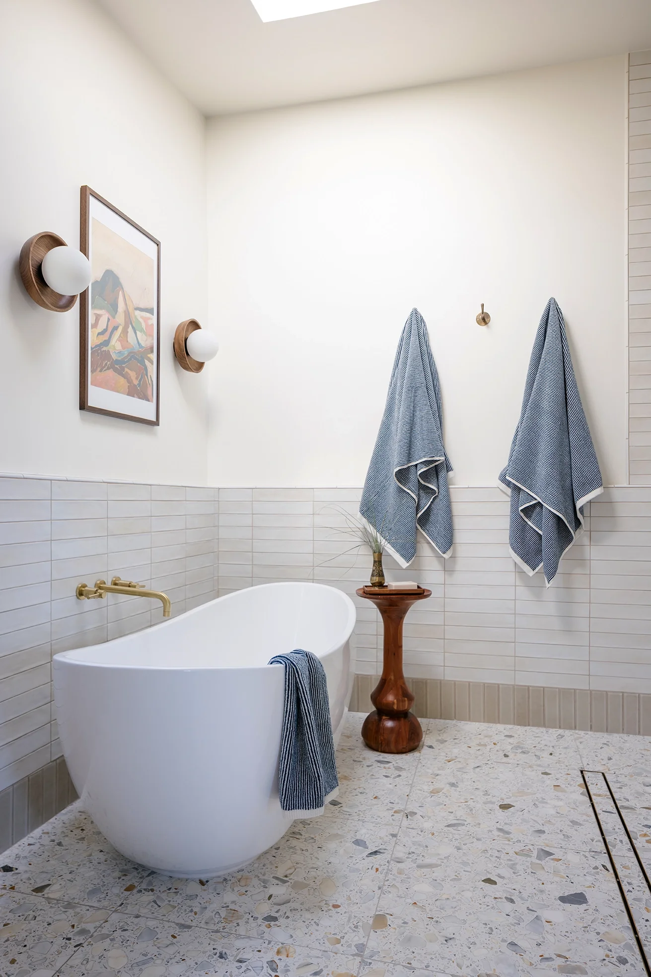 A modern bathroom with a white freestanding bathtub, two blue and white towels hanging on hooks, a small wooden side table with a decorative vase, framed art print, and wall-mounted light fixtures.