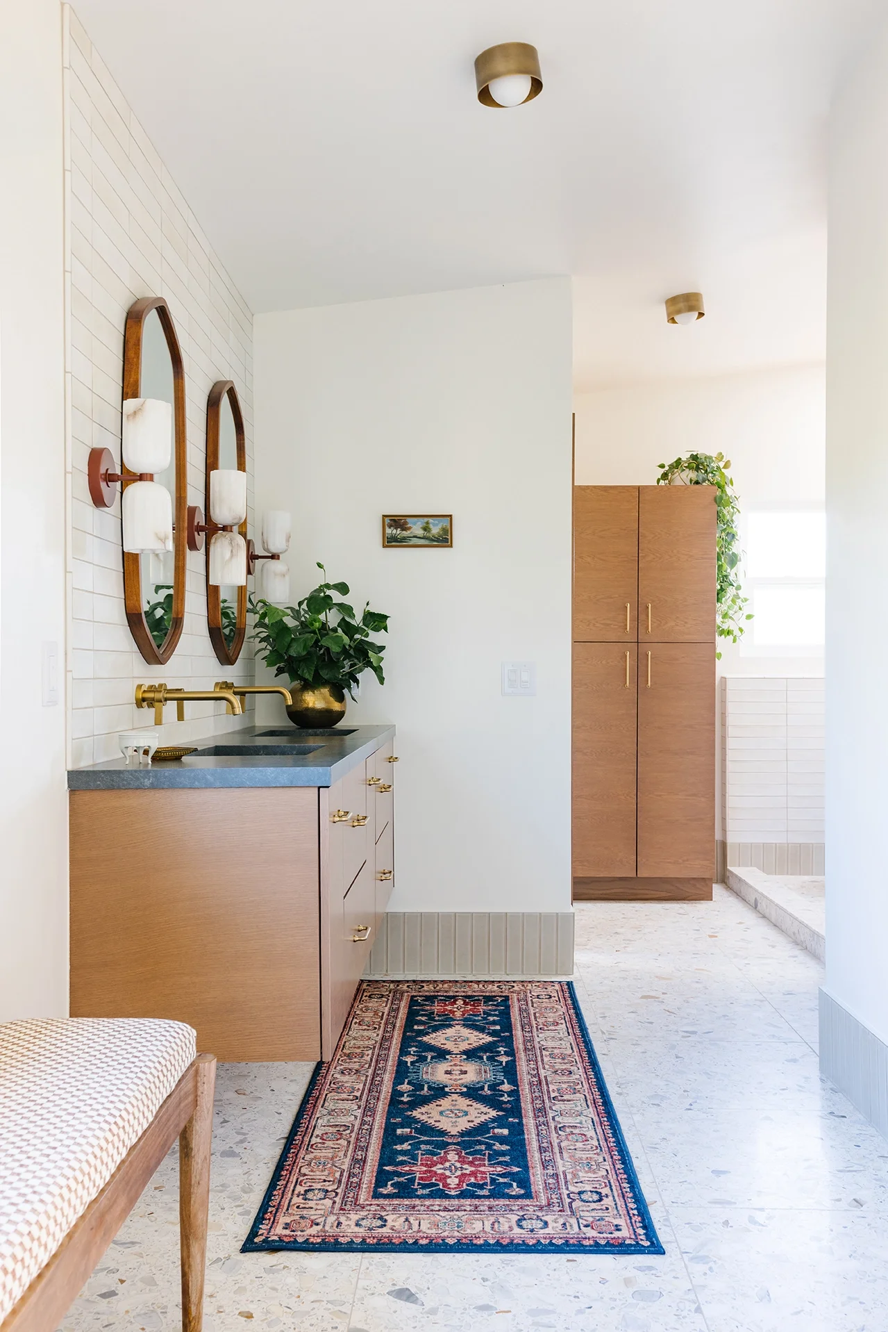 A hallway with a wooden cabinet, a plant, a small framed picture, a colorful patterned rug, a bench, and two oval mirrors with brass and white light fixtures.