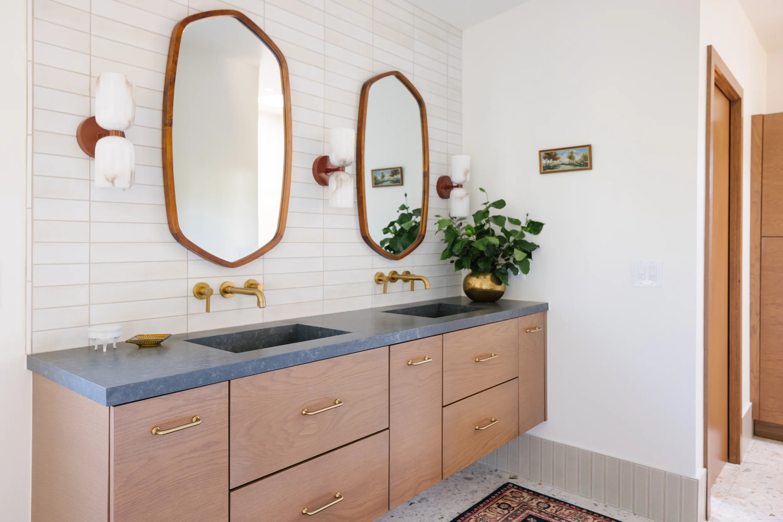 Bathroom vanity with two oval mirrors, gold fixtures, a dark gray countertop, a wooden cabinet with gold handles, a potted plant, and wall sconces with white shades.