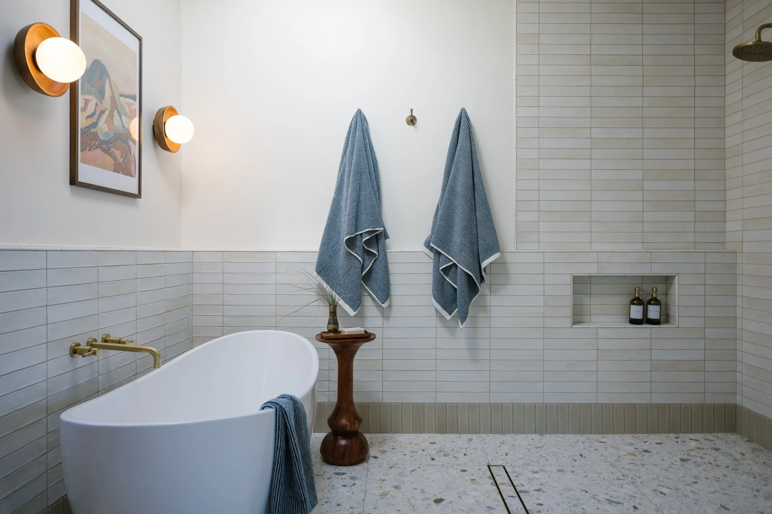 Modern bathroom with white freestanding bathtub, two blue towels hanging on hooks, a small wooden table with a vase and decorative branch, tiled walls and floor, wall-mounted lights, and a built-in shower niche with bottles.