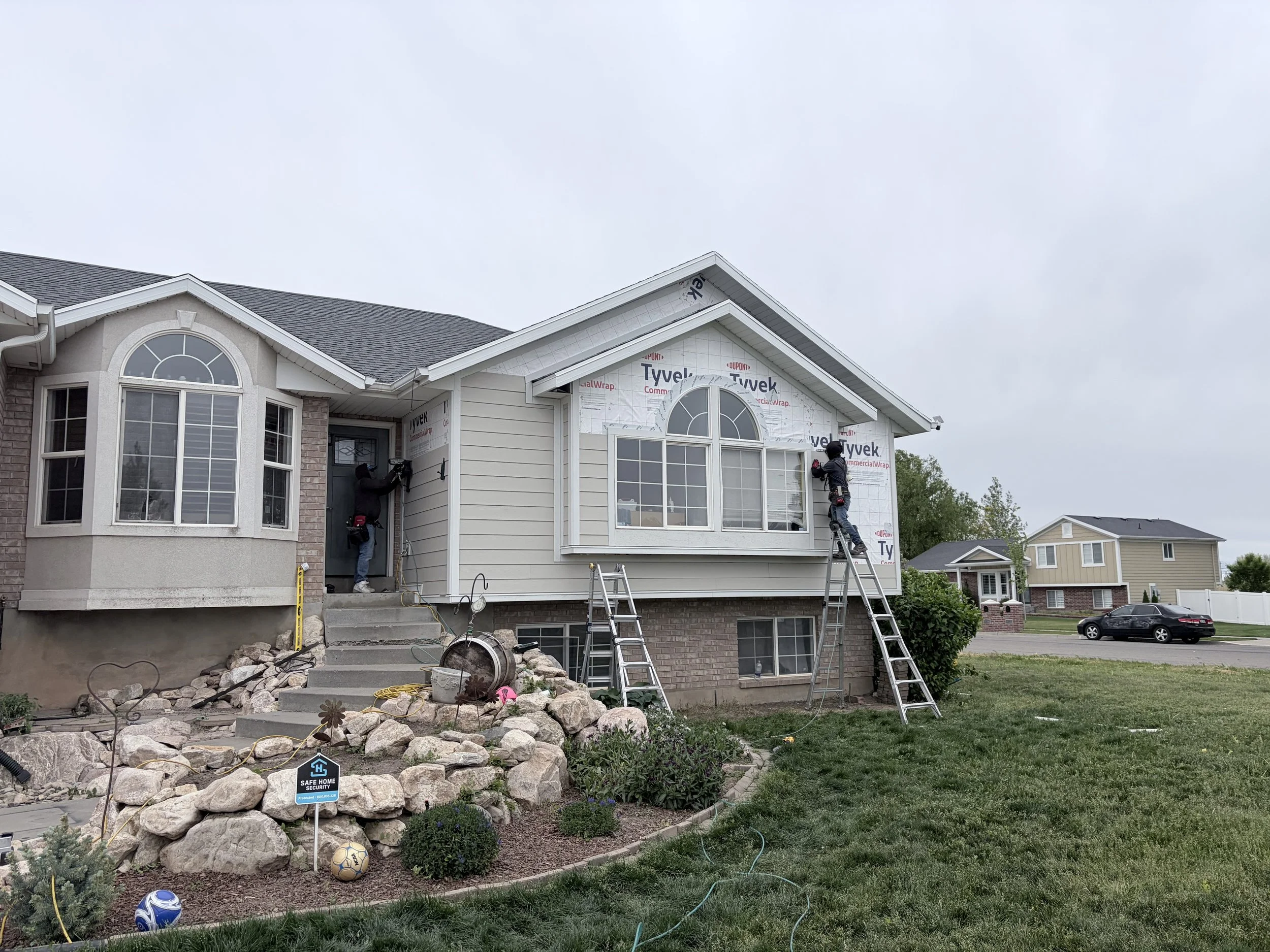 Two construction workers installing siding on the upper part of a house under an overcast sky. The house has large arched windows and a brick foundation. There are ladders, tools, and construction materials around, and a landscaped front yard with small plants and rocks.