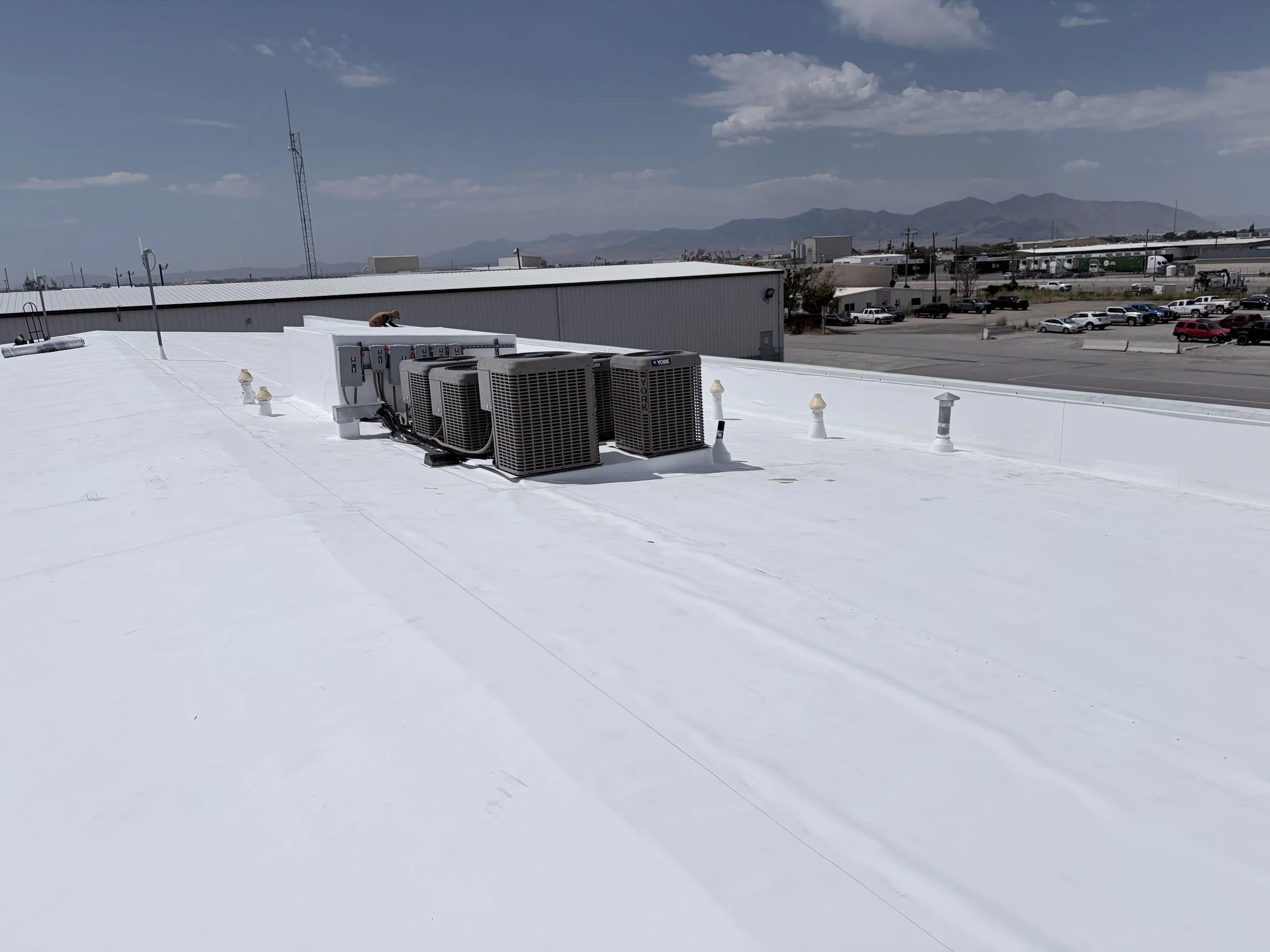 Rooftop with HVAC units, vents, and a worker repairing or installing equipment, with a parking lot, industrial buildings, and mountains in the background under a partly cloudy sky.
