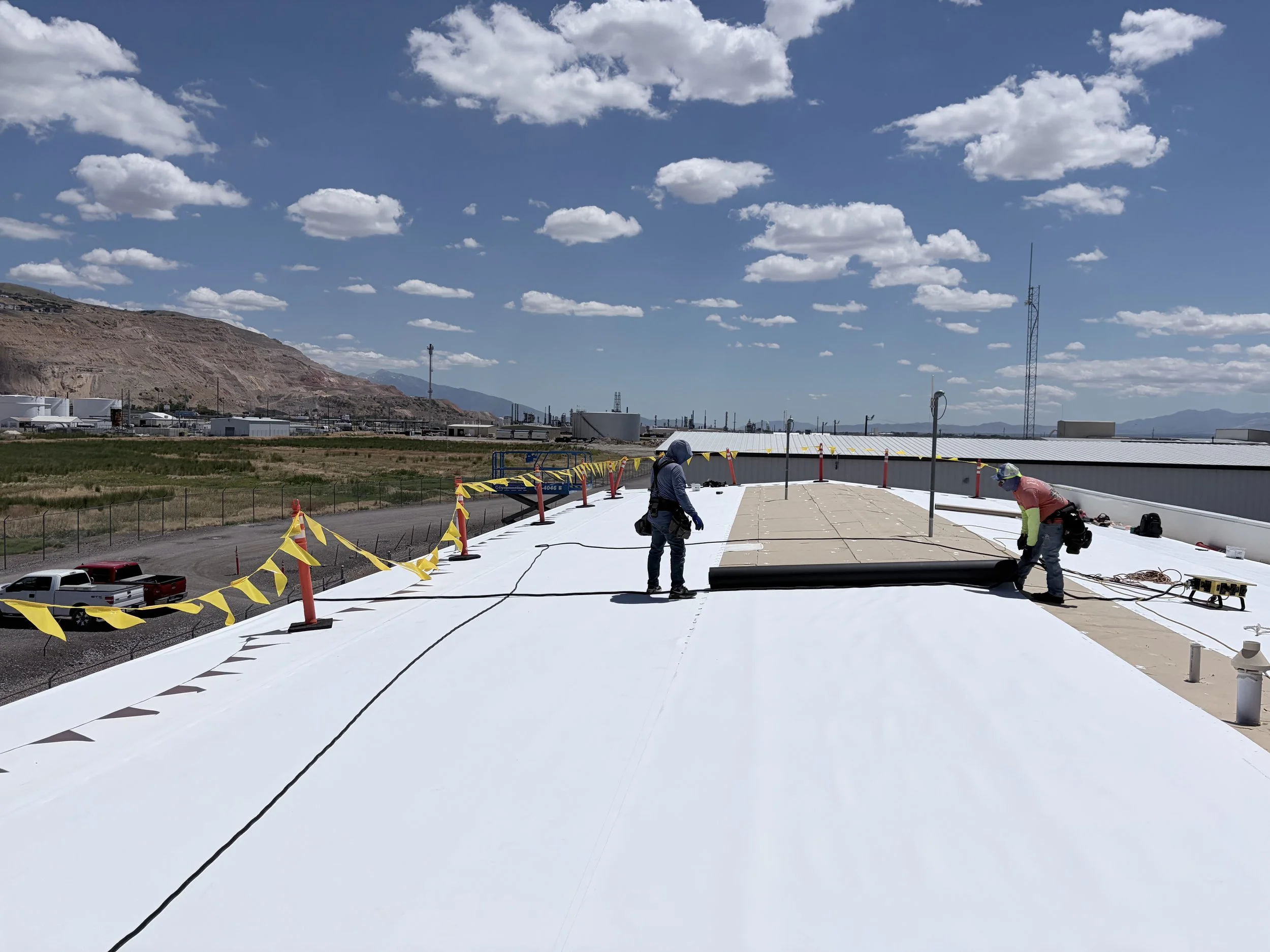 Construction workers installing a white roofing material on a flat rooftop under a partly cloudy sky with mountains in the distance.