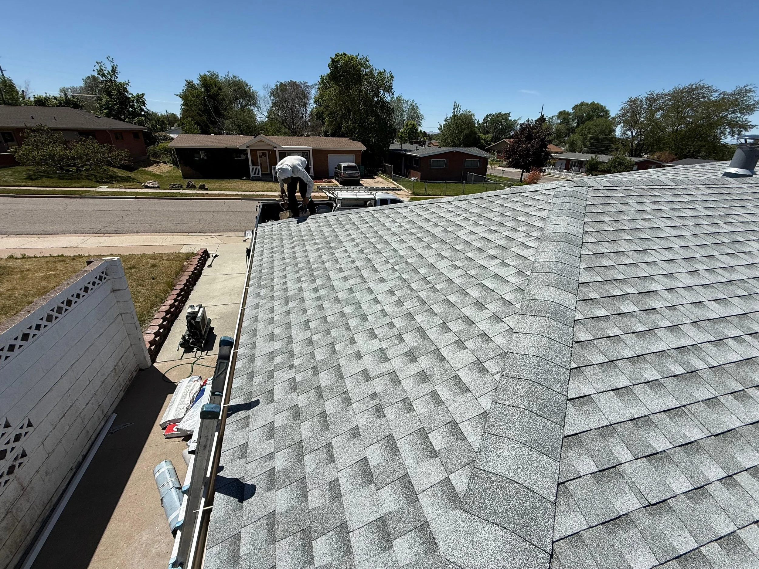 View from a roof showing grey shingles, a worker in white working on the roof, and the street below with houses, trees, and parked cars on a sunny day.