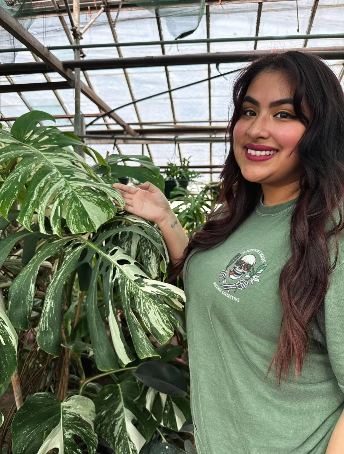 Woman standing in a greenhouse wearing the green “Rooted in Chaos” Mullinix Collective tee, smiling beside a large variegated monstera plant.