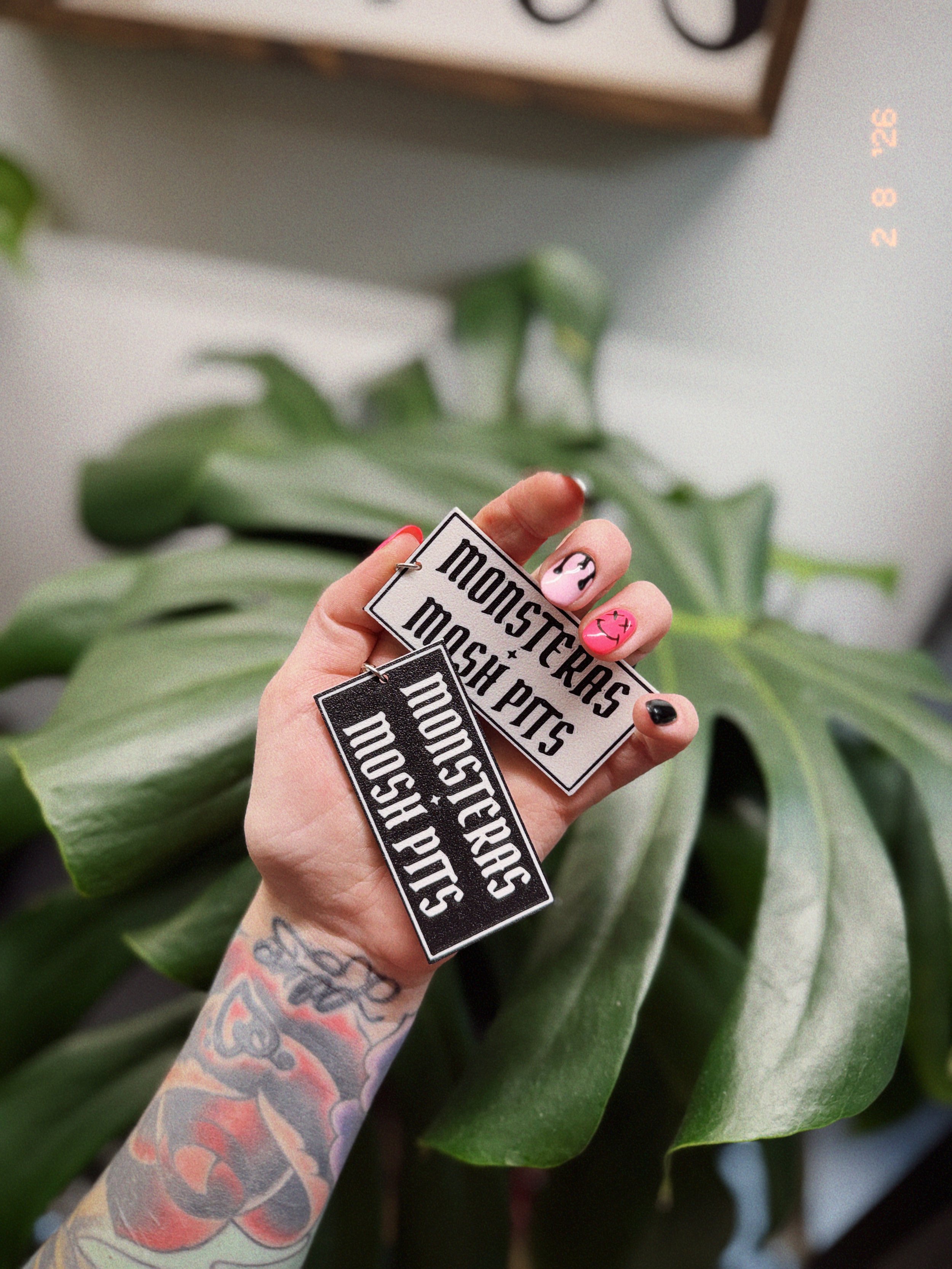 Monsteras + Mosh Pits 3D printed keychains held in hand in front of a large Monstera plant leaf.