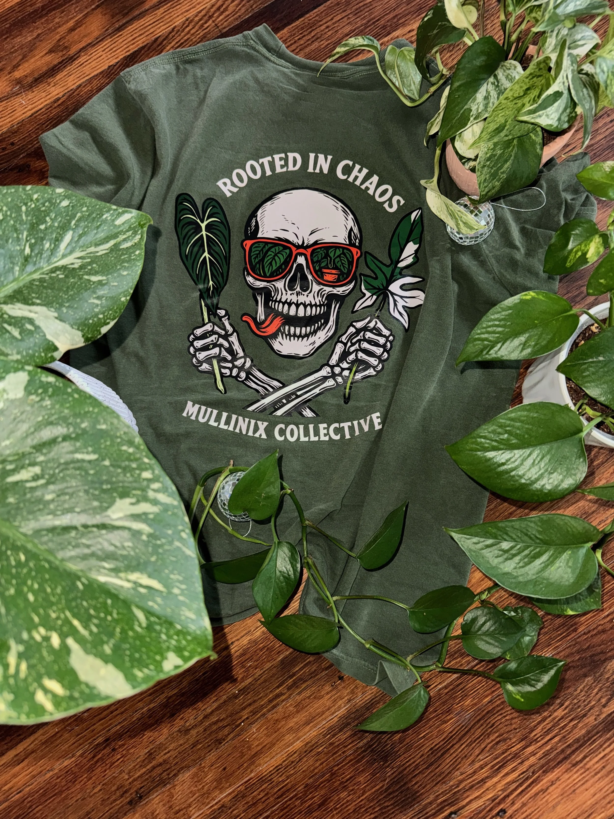 Green “Rooted in Chaos” Mullinix Collective t-shirt laid flat on wood surrounded by pothos and variegated houseplants, showcasing the skull and foliage design.
