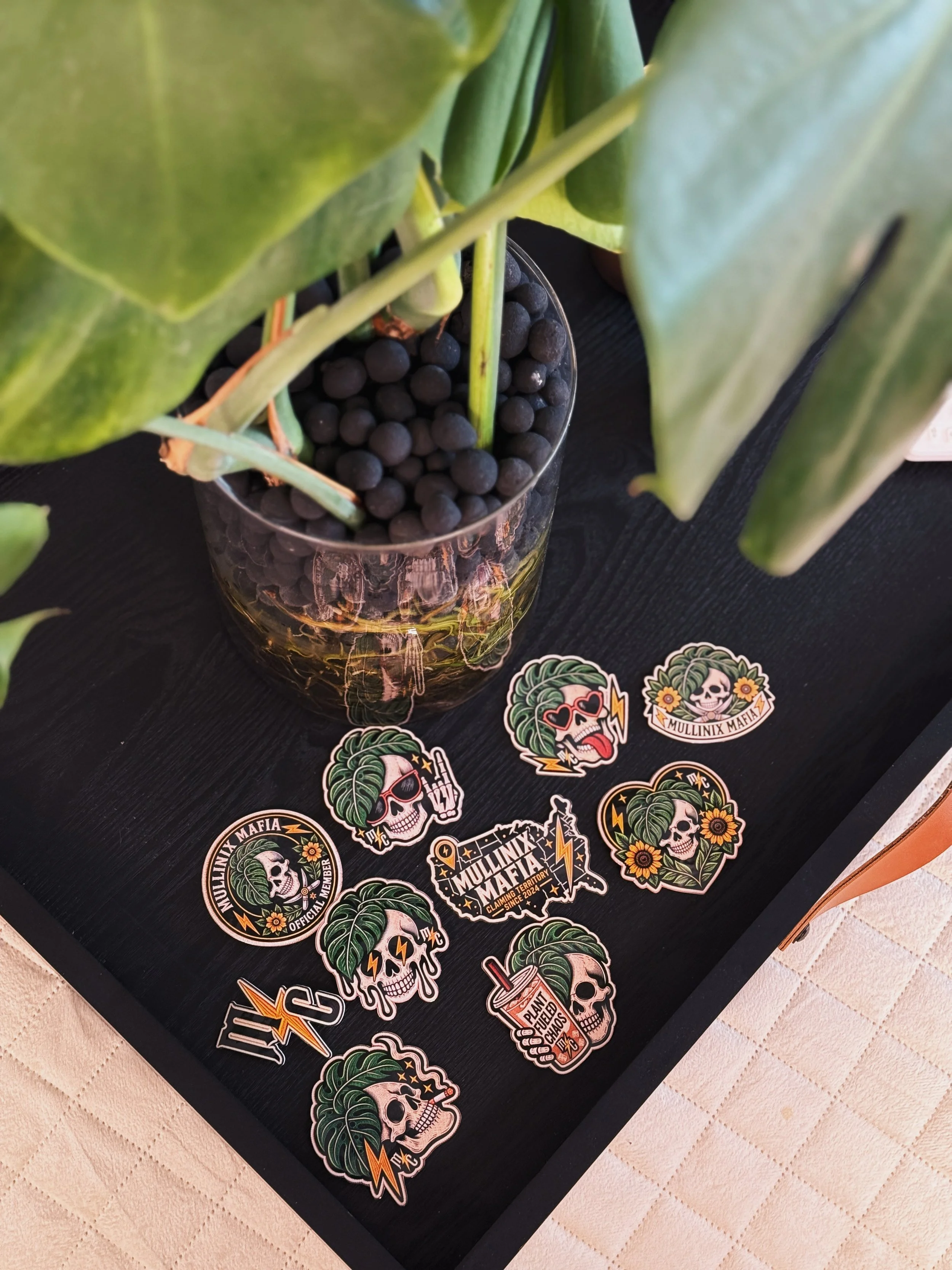 Ten Mullinix Mafia vinyl stickers displayed beside a houseplant featuring skull, monstera and alternative plant designs
