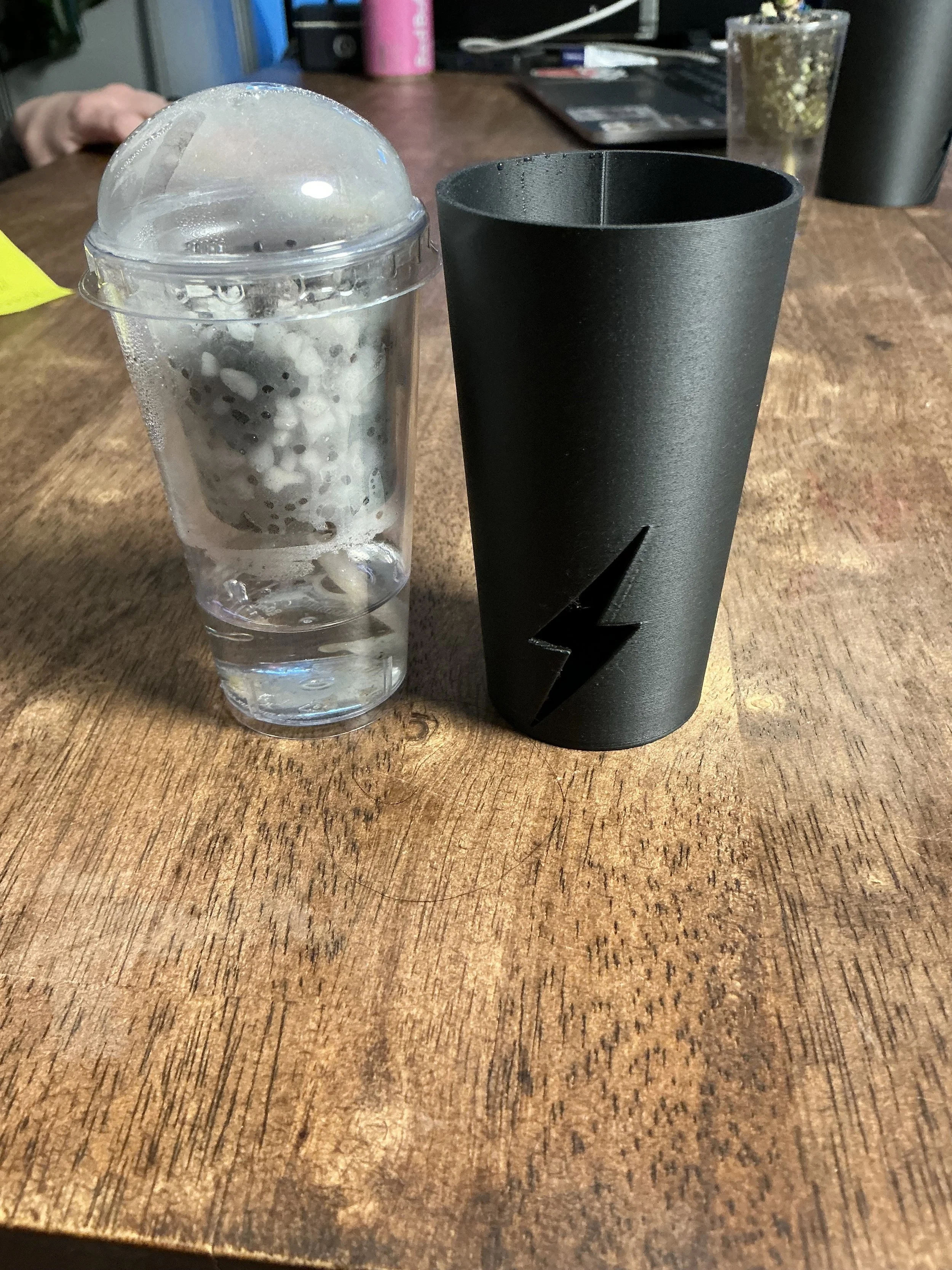 DLux 3oz propagation cup with lid next to black corm cup cover on wooden table for size comparison.