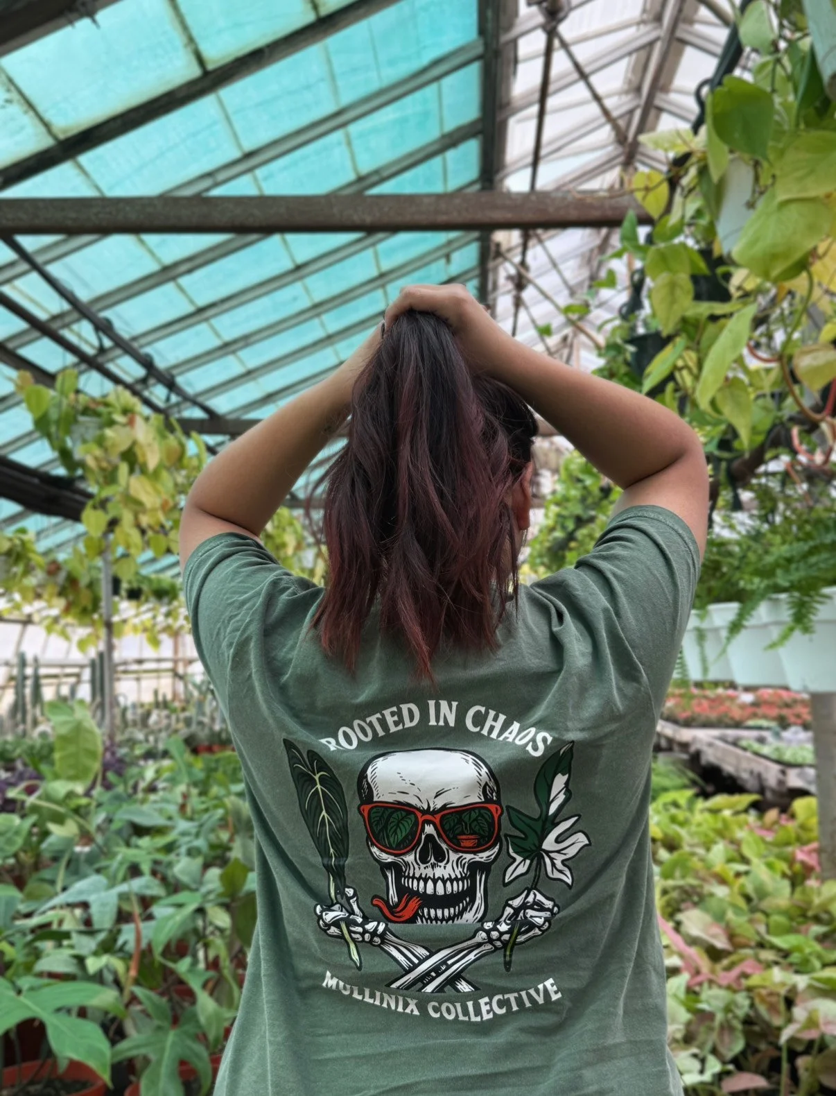 Back view of the green “Rooted in Chaos” Mullinix Collective t-shirt worn in a greenhouse, showing the skull and plant graphic design.
