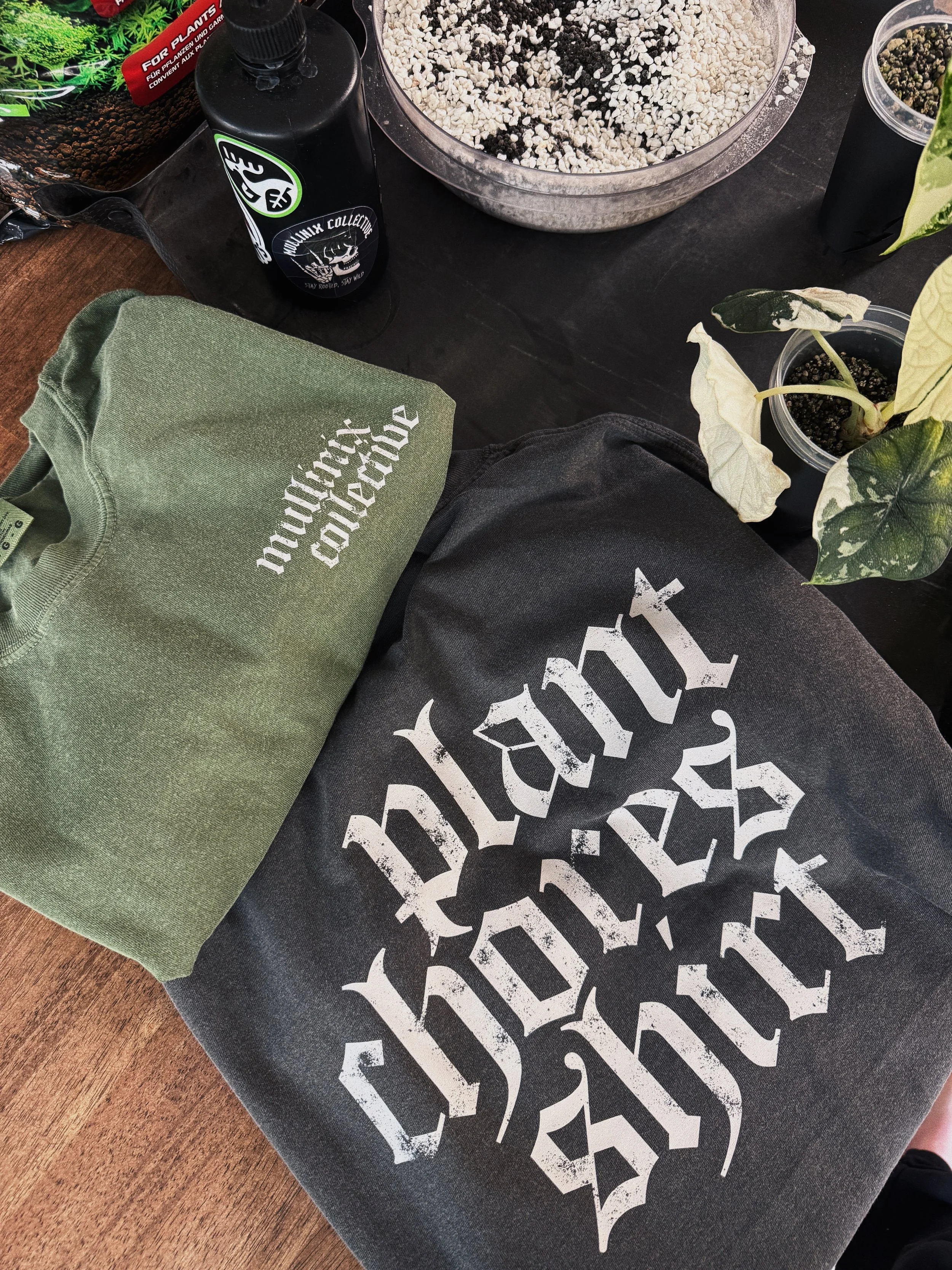Comfort Colors Plant Chores graphic tee with Mullinix Collective logo displayed alongside houseplants and potting supplies